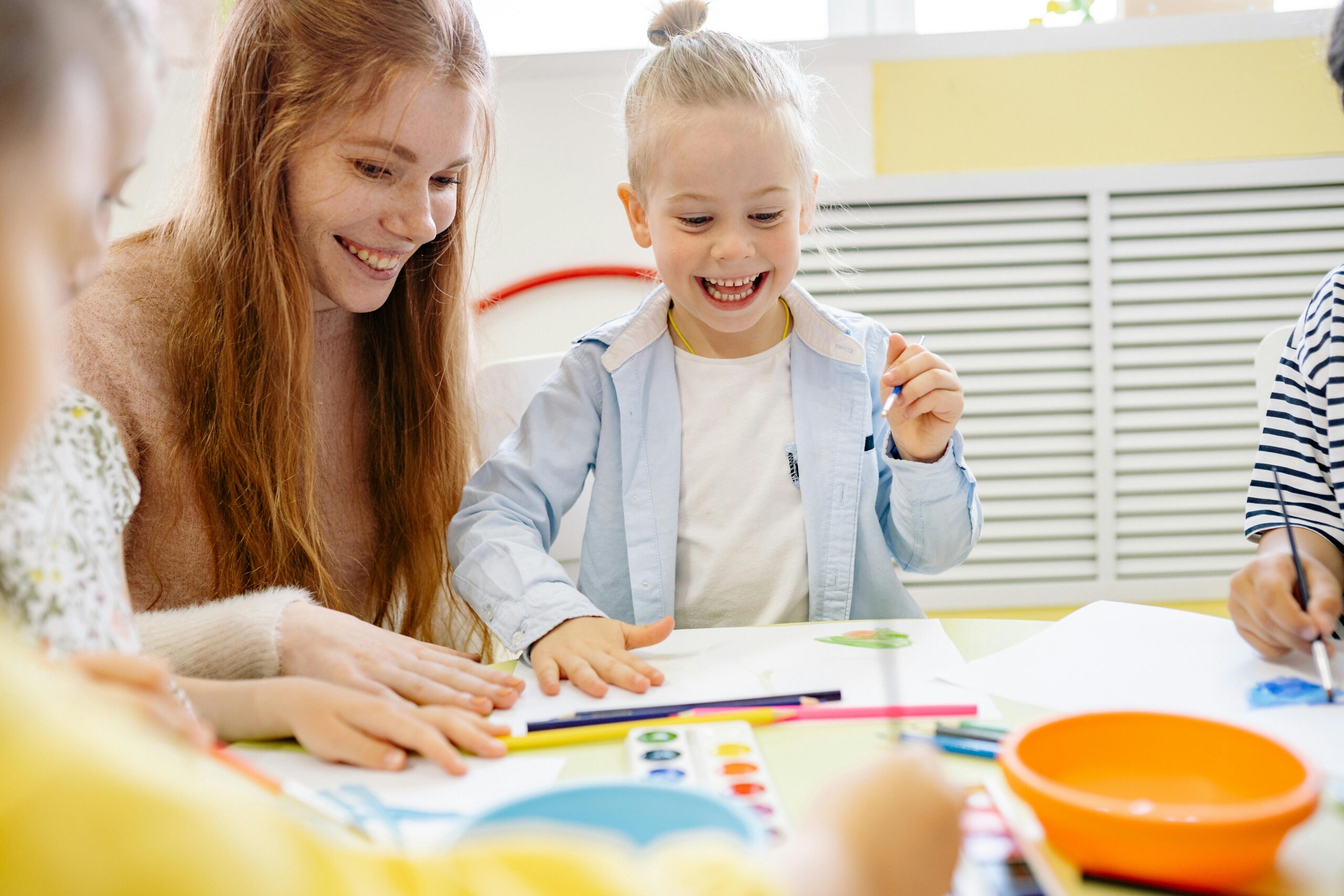 blonde child smiles as teacher does an activity at a table with her