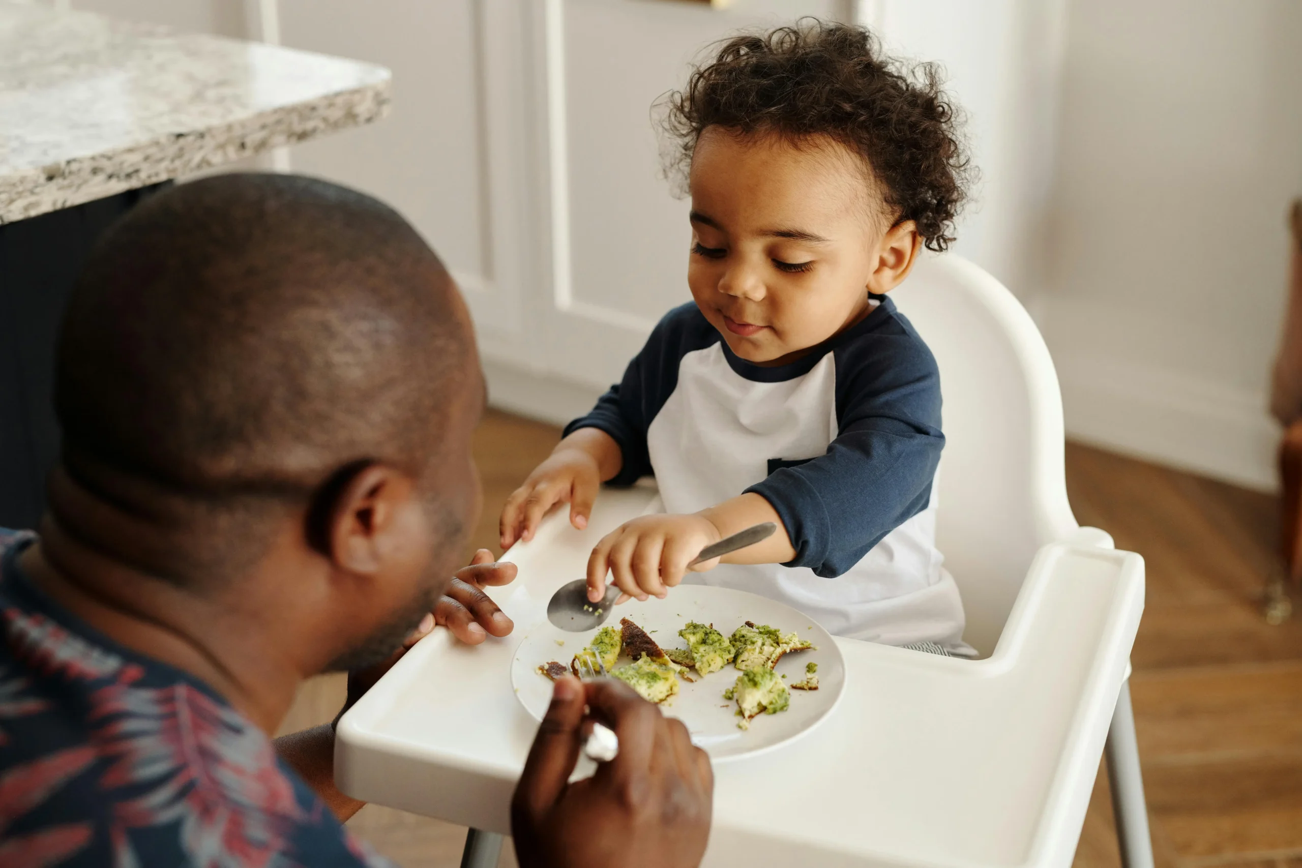 father feeds toddler at high chair during mealtime