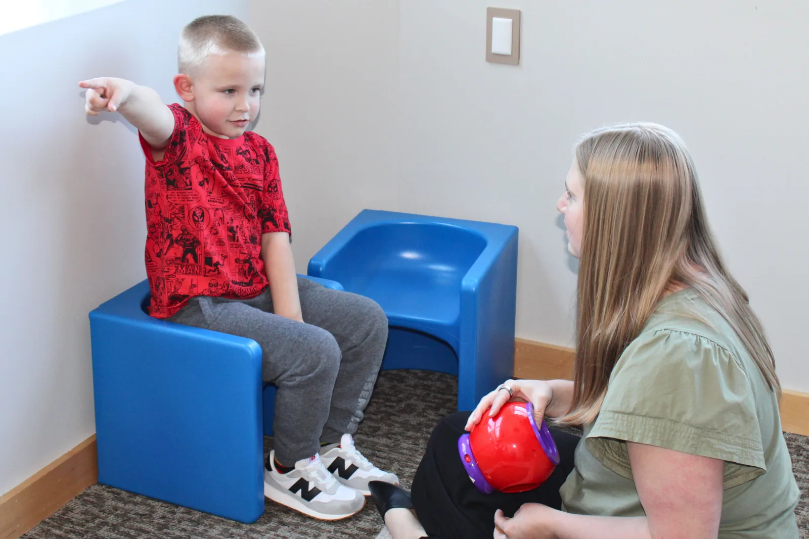 how-to-help-your-child-manage-big-emotions-and-anxiety Young boy seated on a cube chair pointing to a feelings choice while a therapist supports him
